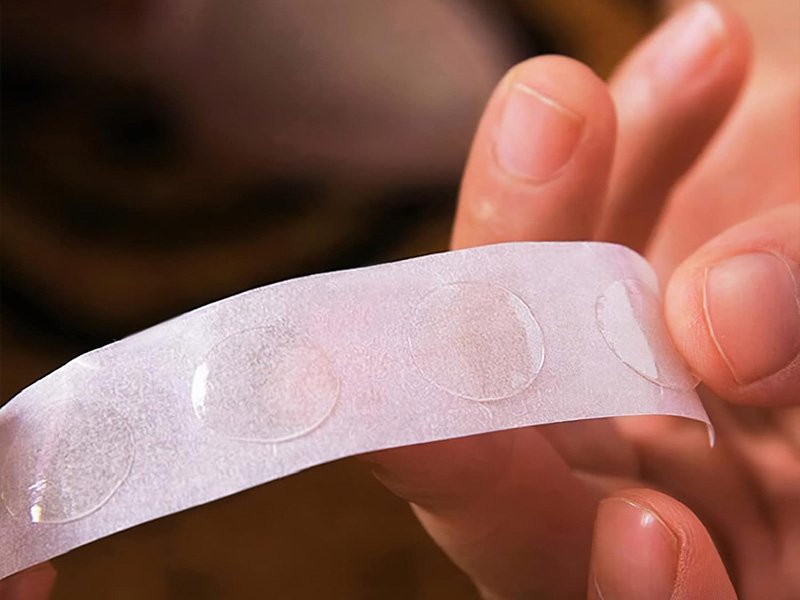 Rouleau de pastilles adhésives double face pour fixer des ballons, tenu entre deux doigts